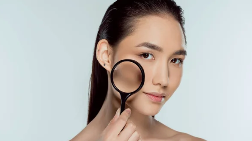一位年輕的亞洲女性將放大鏡靠近臉頰檢查自己的皮膚，在淺藍色背景下表情中性地直視鏡頭。